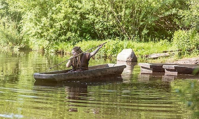 En kvinna i bruna skinnkläder och stort hår sitter i en stockbåt, siktar med ljustret ner i vattnet. vid stranden ligger ytterligare tre stockbåtar